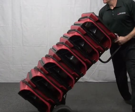 Person pushing a stack of Phoenix AirMax Radial Air Movers on a dolley against a white wall.