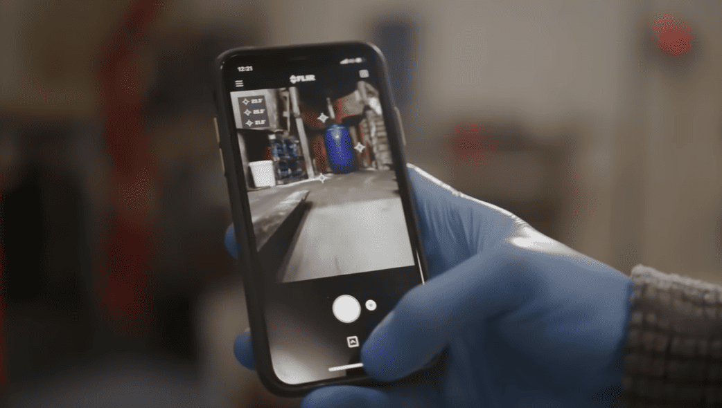 Hand wearing a blue glove holding a smartphone FLIR ONE Edge Pro thermal camera with a blurred indoor background