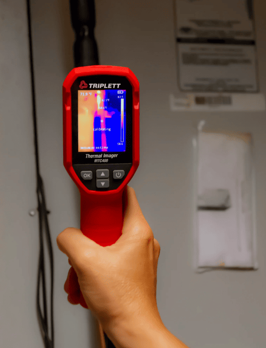 Person holding a red IRTC400 thermal imaging device labeled 'Triplett' against a wall.