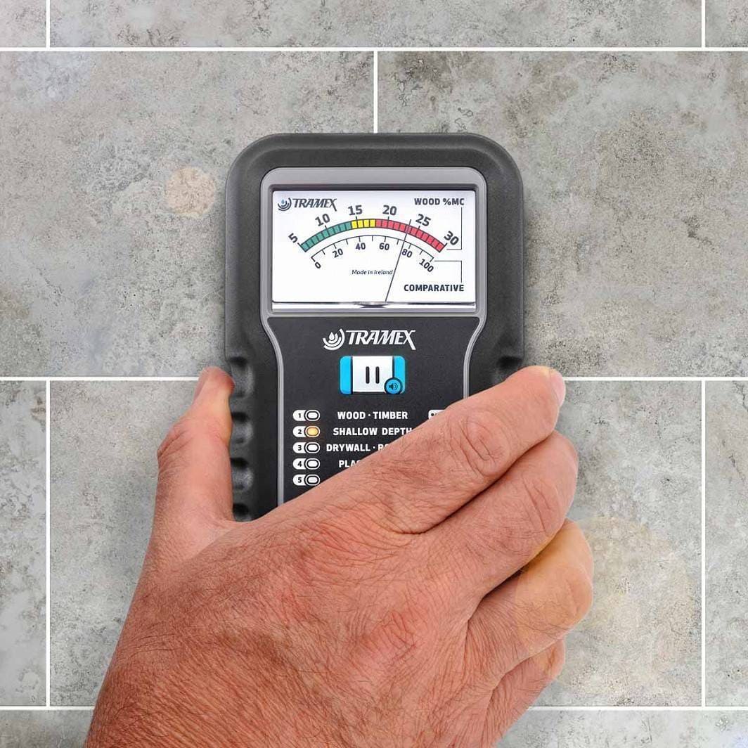 Hand holding a Tramex ME5 moisture meter against a tiled wall background