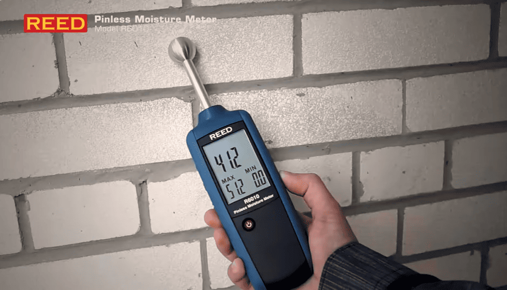 Hand holding a REED_R6010 moisture meter against a brick wall.