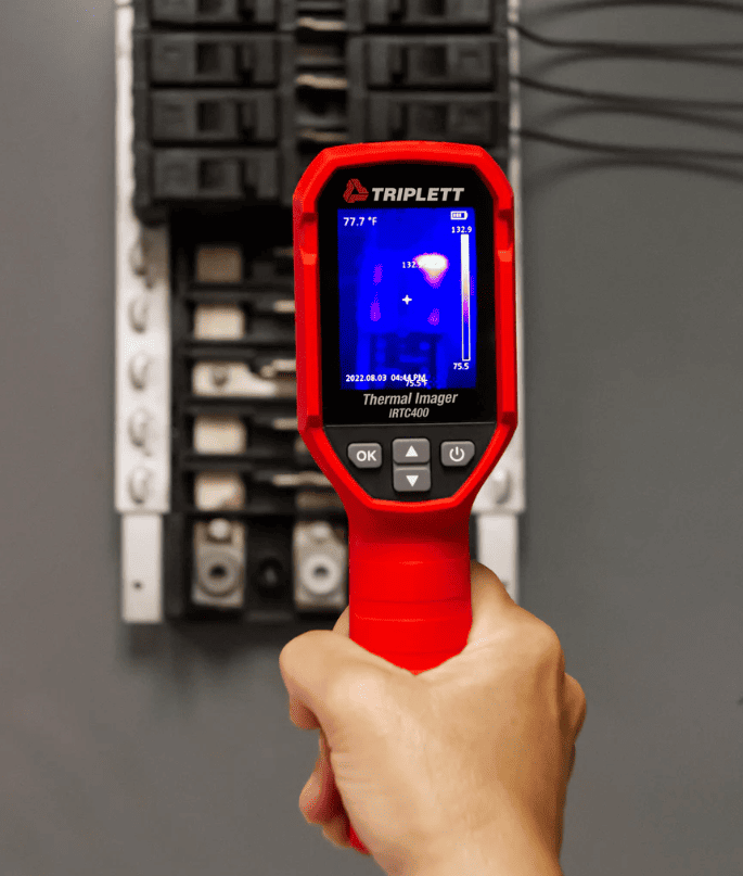 Hand holding a red Triplett IRTC400 thermal imager in front of an electrical panel.