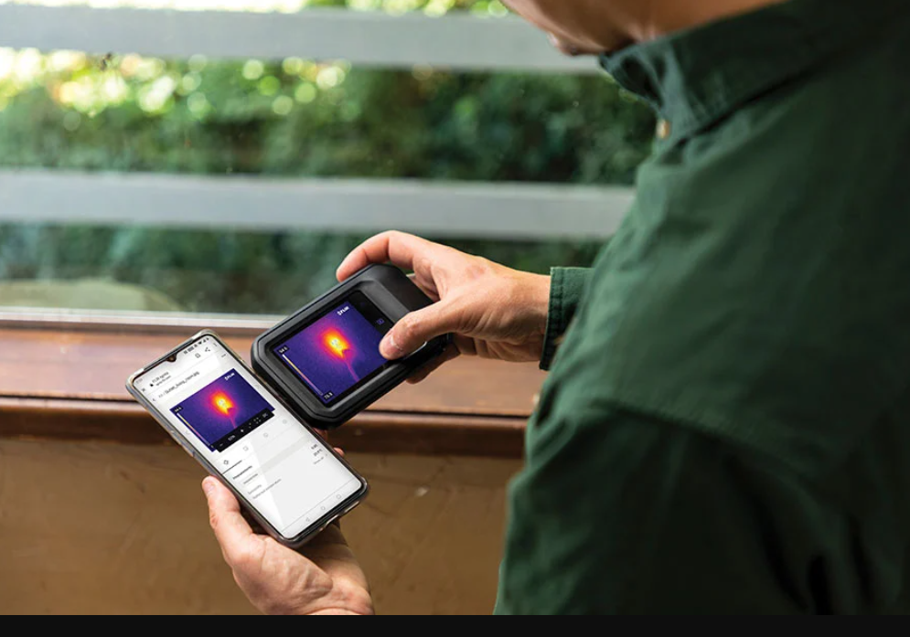 Person holding a smartphone and a flir c3 x thermal imaging device with app interface on screens.