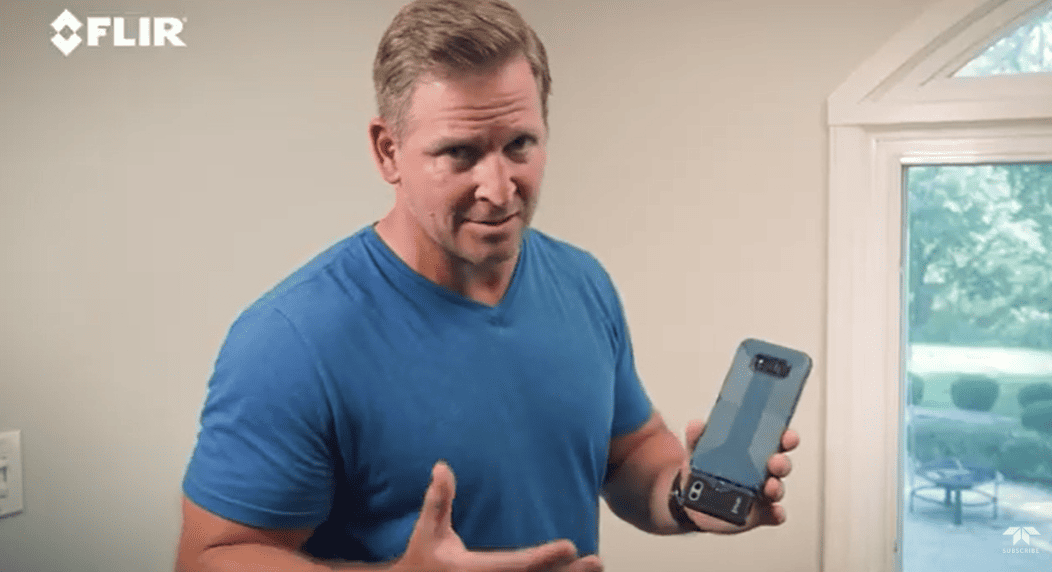Man holding a FLIR thermal imaging device in a room with a window showing outdoor view.