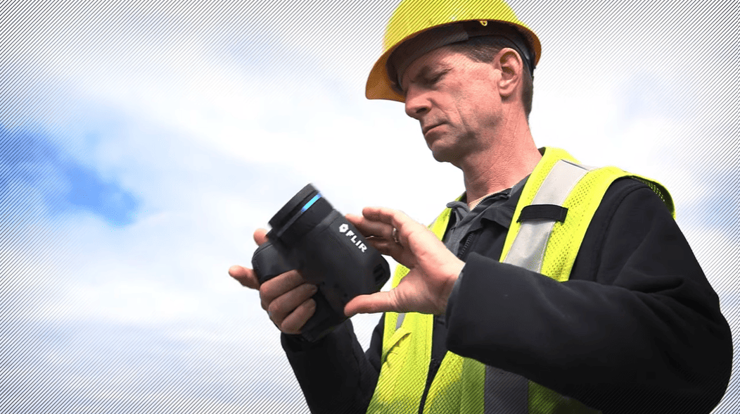 Person in safety gear holding a flir t530 thermal camera with a blurred sky background