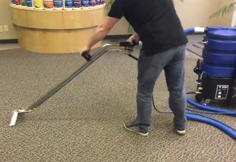 Person using a ninja carpet extractor 500 psi with heat on a carpeted floor with cleaning equipment in the background.