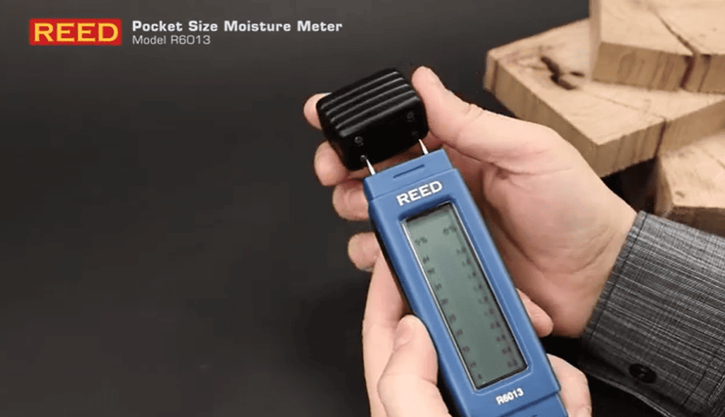 Hand holding a REED r6013 pocket size moisture meter with wooden blocks in the background