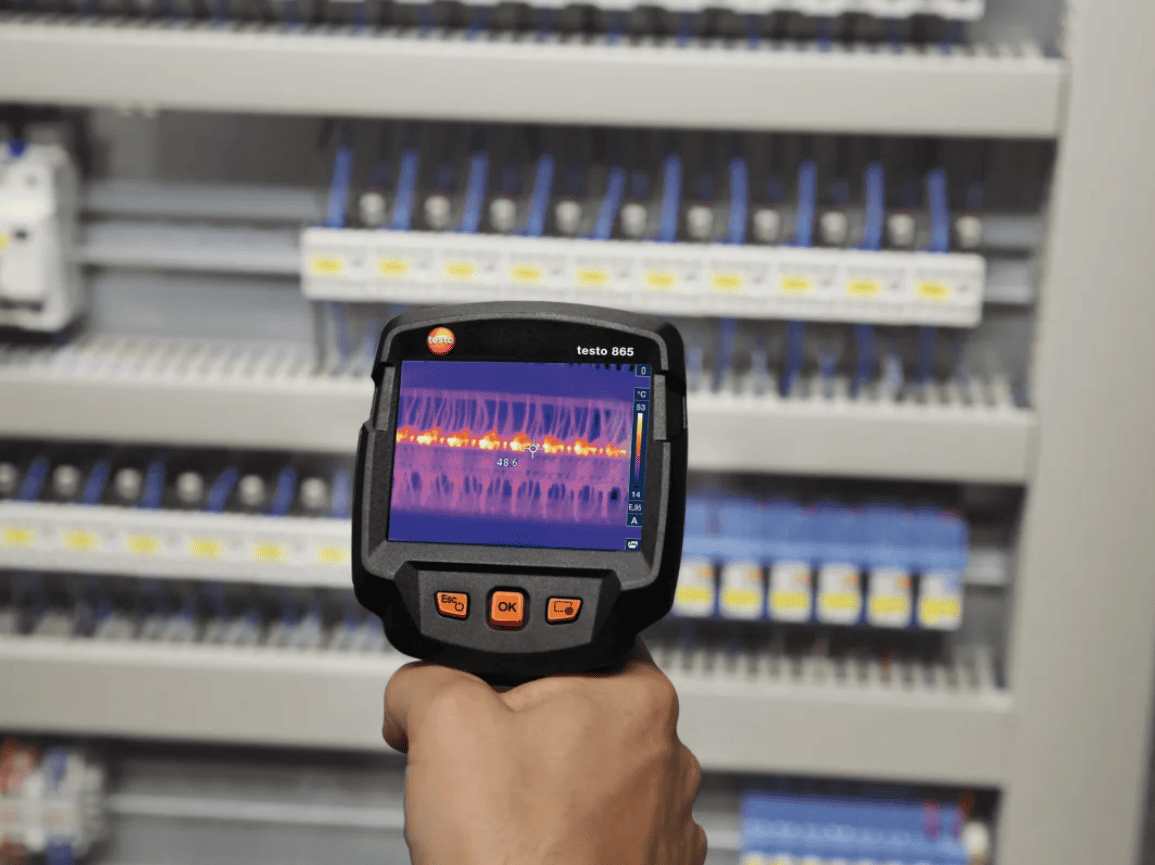 Hand holding a testo 865s thermal imager in front of electrical panels.