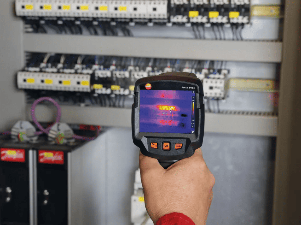 Hand holding a testo 868s thermal imaging device in front of an electrical panel.