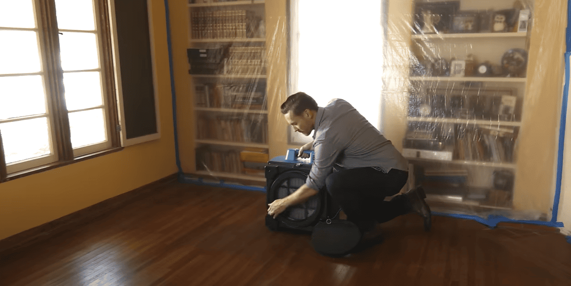 Person kneeling on a wooden floor with a x3580, surrounded by plastic-wrapped furniture.
