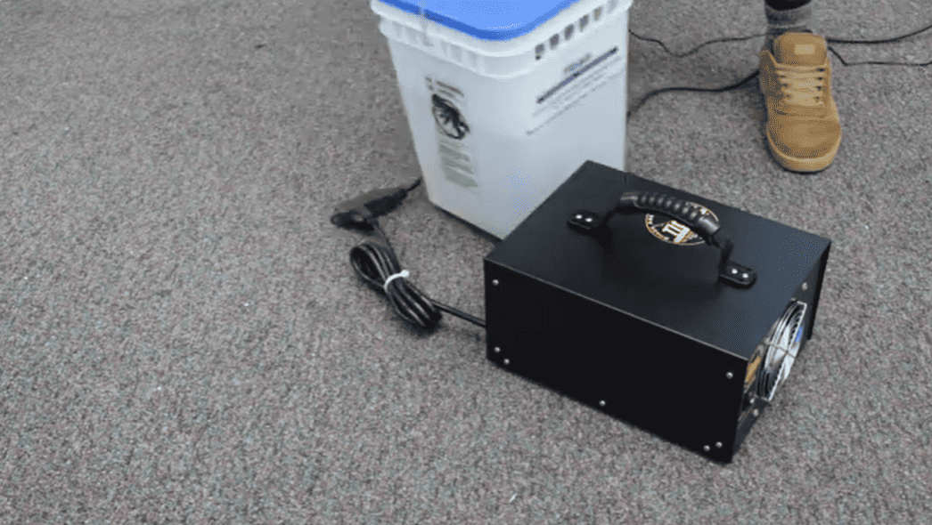 Black Hydroxyl Generator on a gray floor next to a white container and person's foot.