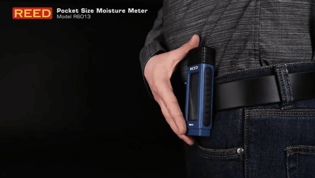 Hand holding a blue REED r6013 pocket-size moisture meter against a dark background