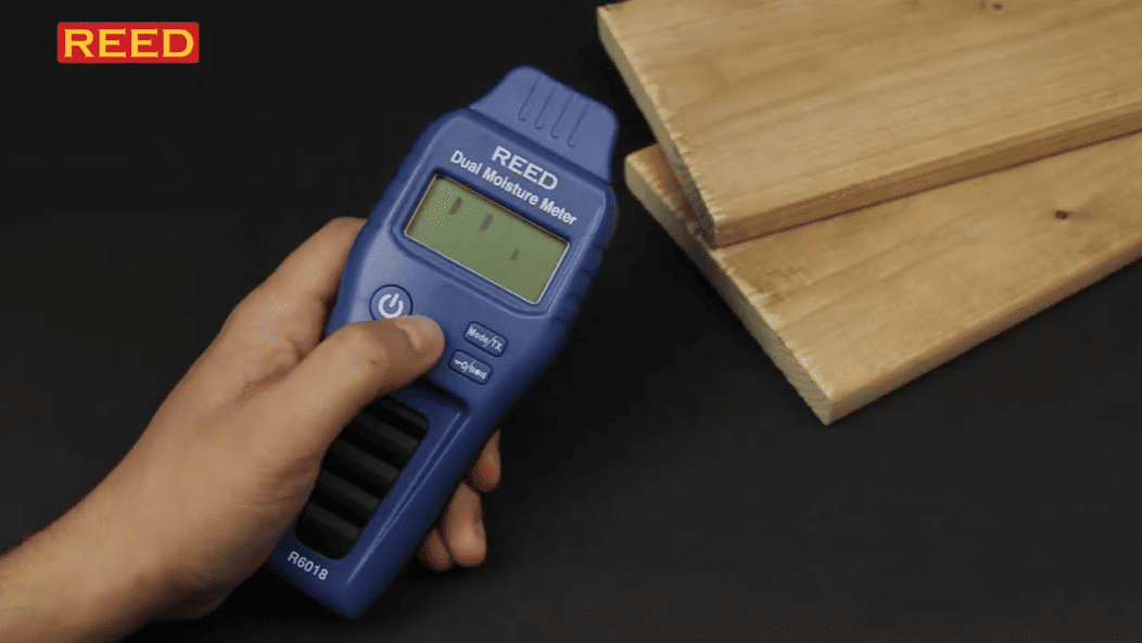 Hand holding a blue REED moisture meter next to wooden planks on a dark background