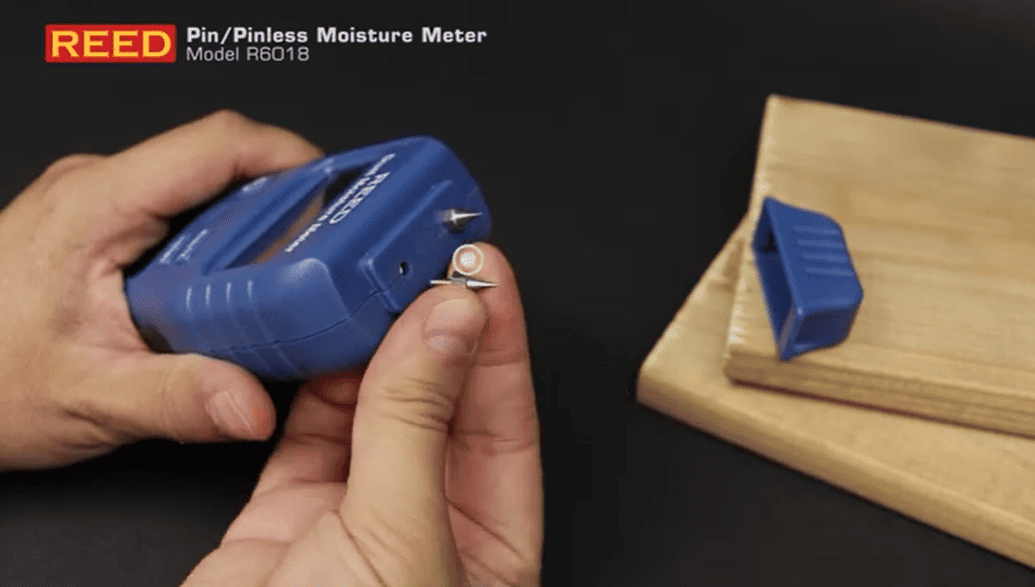 REED R6018-KIT Hand holding a blue REED pinless moisture meter on a dark background