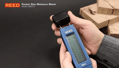 Hand holding a REED r6013 pocket size moisture meter with wooden blocks in the background