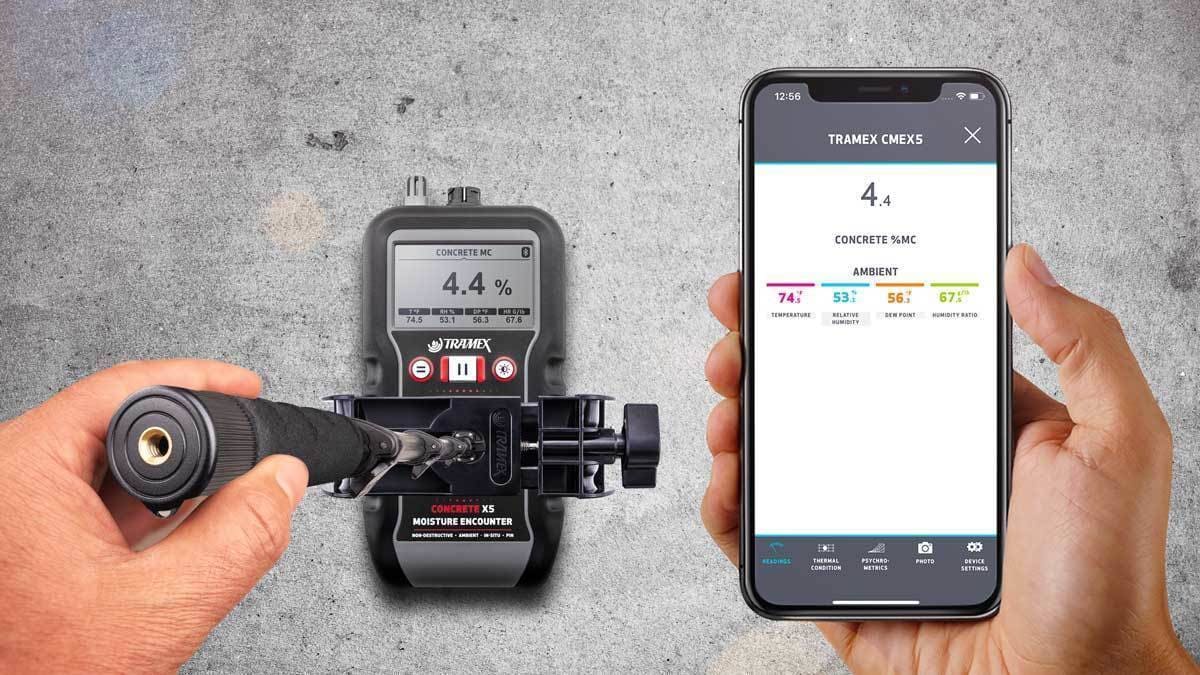 Hand holding a tramex x5 moisture meter and smartphone displaying Tramex software on a gray background